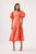 Woman wearing coral Maia Off Shoulder Midi Dress from Aje featuring relaxed skirt with curved panel seams and assymetric neckline adorned with short puff sleeves.