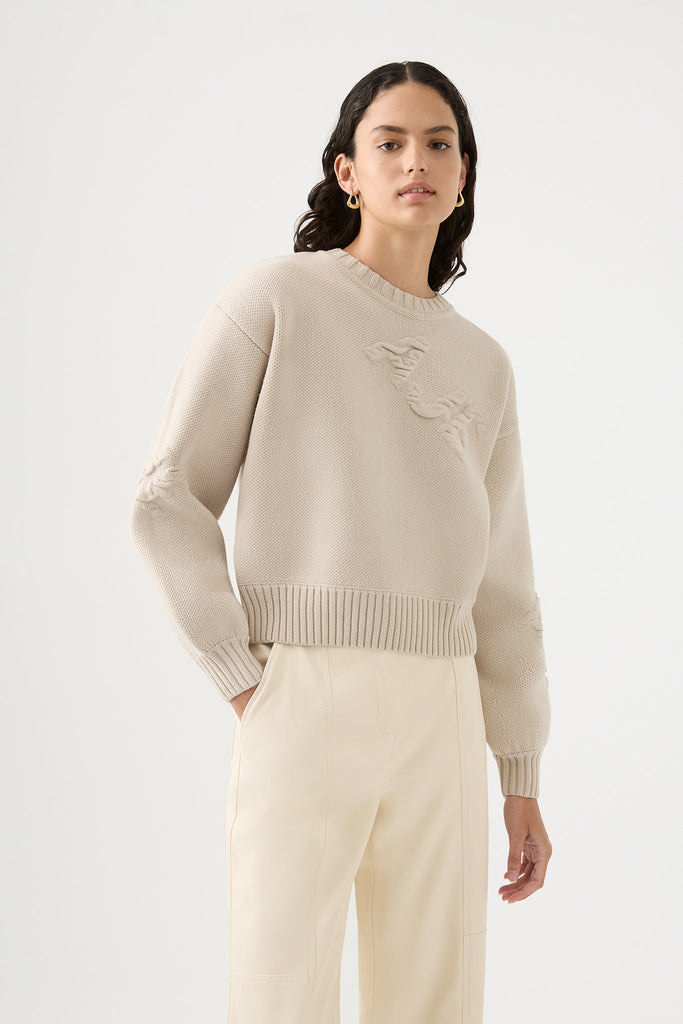 Woman wearing Superlative Logo Jumper by Aje in beige designed with a relaxed fit, featuring blouson sleeves, crew neck, and ribbed cuffs.