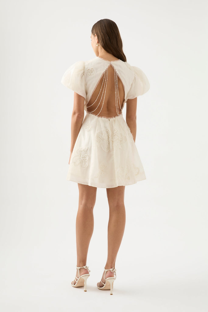 Woman wearing Vera Beaded Flower Mini White Dress from Aje with dropped shoulders with gathered sleeves, adorned with hand-beaded pearls, featuring cut out back with pearl trim.